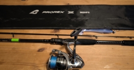 Spinnrute Daiwa Prorex X Spin mit daran befestiger Angelrolle Shimano Nasci 4000XG. Daneben liegen ein blauer Gummifisch mit Jigkopf und ein Rutenfutteral