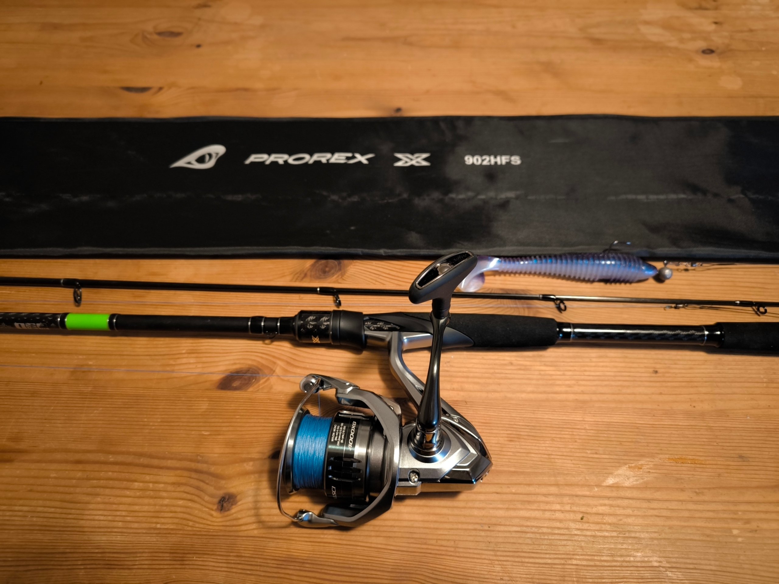 Spinnrute Daiwa Prorex X Spin mit daran befestiger Angelrolle Shimano Nasci 4000XG. Daneben liegen ein blauer Gummifisch mit Jigkopf und ein Rutenfutteral